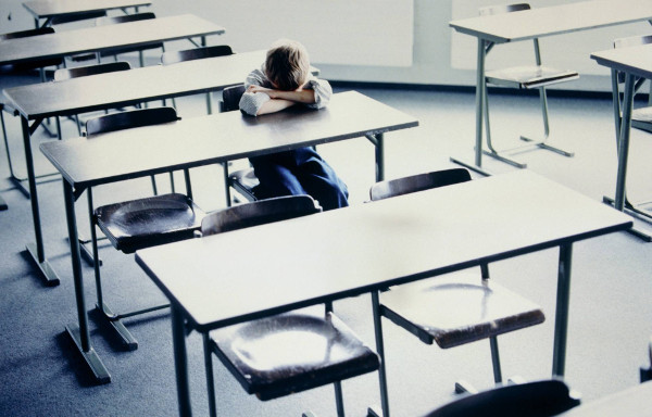 imagen de un niño con la cabeza entre los brazos sentado solo en una mesa de una clase
