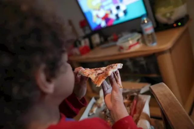 Niño/a come un trozo de pizza delante de la televisión